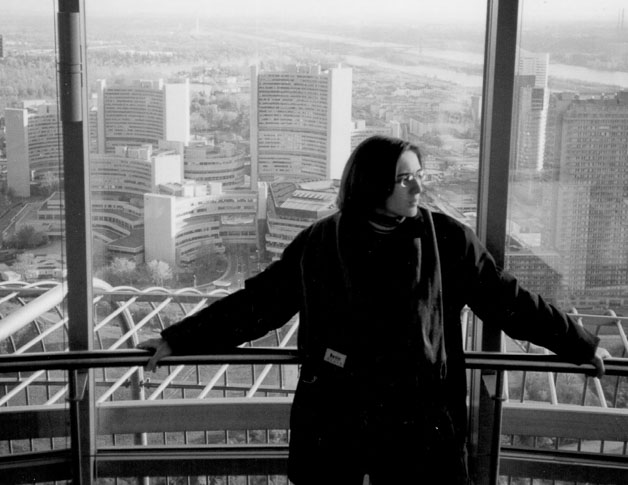 Mlada žena s naočalama ispred staklene stijene, gleda u stranu. Ispod nje panoramski pogled na grad.