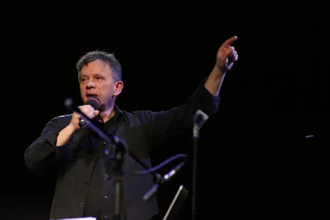 Muškarac s mikrofonom u ruci. Njegova druga ruka pokazuje prema gore.