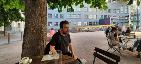 Ein junger Mann mit Bart sitzt an einem Tisch in einem Biergarten. Hinter ihm sind ein großer Platz und ein Kinogebäude.