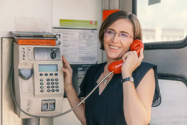 Nasmejana žena sa naočarima u crnoj haljini razgovara telefonom u staroj telefonskoj govornici.